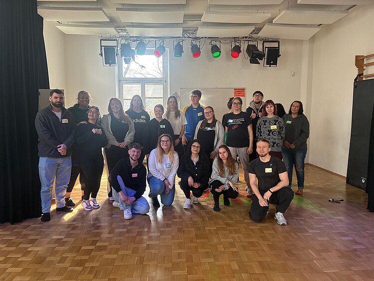 Gruppenfoto von Teilnehmenden und Betreuungspersonen in einem großen Probenraum mit Parkettboden und farbiger Bühnenbeleuchtung.