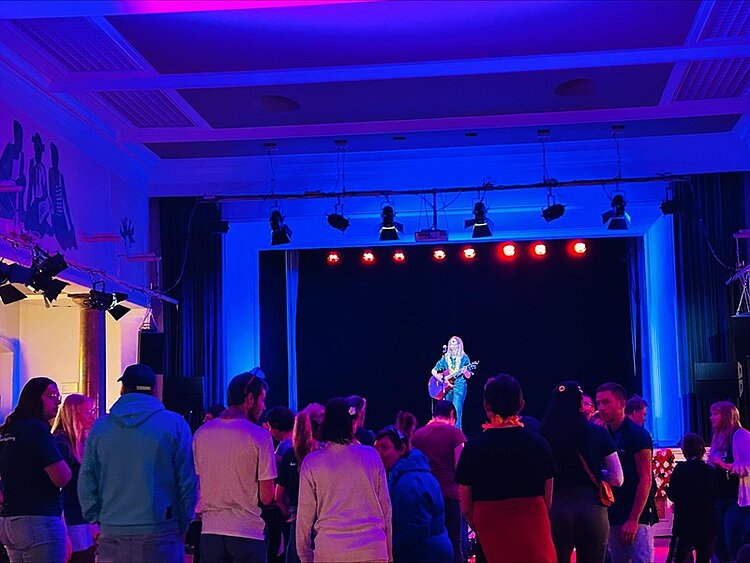 Eine Person steht auf einer Bühne und spielt Gitarre. Die Bühne ist mit roten und blauen Lichtern beleuchtet. Im Vordergrund steht eine Gruppe von Menschen, die zur Bühne blicken. Die Decke und Wände des Raumes sind ebenfalls in blaues Licht getaucht. An der linken Wand ist ein großes Wandbild mit mehreren Figuren zu sehen.