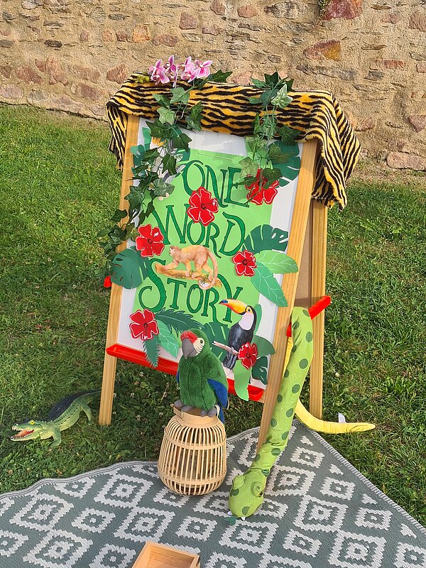 Kennenlerntag der Euro Akademie Rochlitz Eine bemalte Tafel mit der Aufschrift „One Word Story“, dekoriert mit Blättern, Blumen und Tiermotiven. Davor liegen ein Plüschpapagei, ein Stoffkrokodil und eine Stoffschlange als Dschungel-Deko.