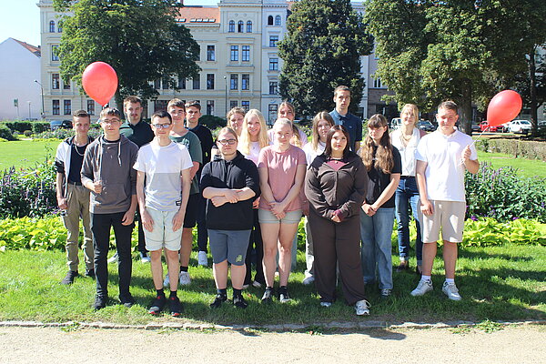 Herzlich Willkommen zum ersten Schuljahr an der Euro Akademie Görlitz! Auf einer sonnigen Wiese inmitten der Stadt haben sich die neuen Auszubildenden für ein Gruppenfoto versammelt. Zwei rote Luftballons haben es auch auf das Bild geschafft.