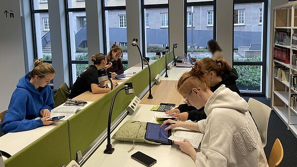 Heller Lernraum mit langen weißen Tischen und grünen Trennwänden. Mehrere Personen in lässiger Kleidung arbeiten konzentriert an Laptops, Tablets und Notizen; im Hintergrund Regale mit Büchern und Ordnern.