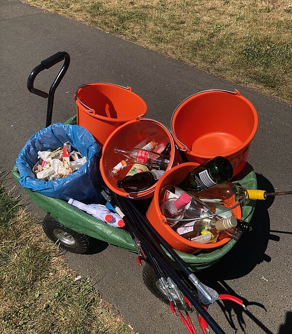 Ein mit Müll gefüllter Bollerwagen steht auf einem Gehweg. Der Wagen enthält mehrere orange Eimer, die teilweise mit leeren Flaschen gefüllt sind und eine blaue Plastiktüte mit Abfall. Auf dem Wagen befinden sich auch mehrere Müllzangen.
