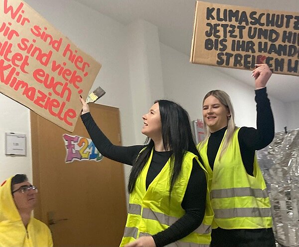 Von Gothic bis Punk! Zwei Azubis in neongelben Warnwesten stellen Fridays For Future Generation dar und halten Demonstrationsplakate hoch.