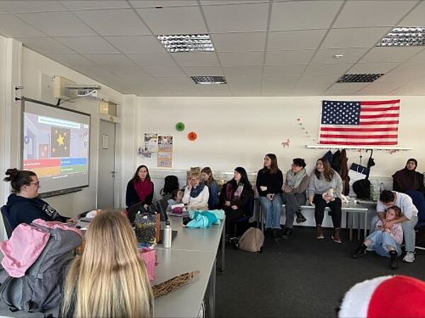 Eine Gruppe von Schülern sitzt in einem Klassenzimmer und schaut aufmerksam auf eine Projektion an der Wand. Einige Schüler sitzen an Tischen, während andere auf dem Boden sitzen. Im Hintergrund hängt eine amerikanische Flagge an der Wand.