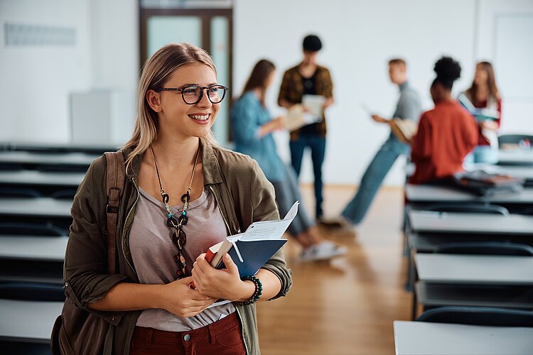 Schulgeldbefreiung 2024/2025 Ein Lehrraum mit Schülerinnen und Schülern in verschiedenen Aktivitäten. Im Vordergrund hält eine junge Frau mit Brille Bücher im Arm. Sie hat eine Tasche über die Schulter gehängt, wie zum Aufbruch oder beim Ankommen. Im Hintergrund sind unscharf andere Schüler*innen stehend oder anlehnend in einer Gruppierung zu sehen. Sie scheinen sich zu unterhalten sich.
