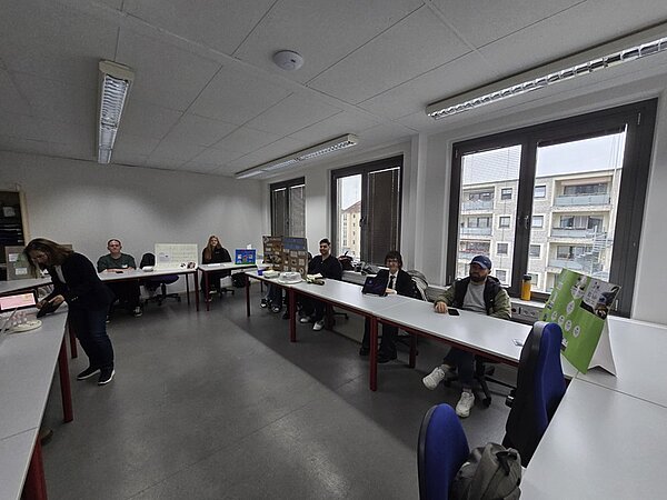 Mehrere Auszubildende sitzen an einem langen Tisch in einem hellen Raum mit großen Fenstern. Auf dem Tisch stehen verschiedene Gegenstände, darunter Kartons, technische Geräte und Papierstapel. Eine Person steht links und beugt sich über den Tisch, während die anderen sitzen und in Richtung Kamera blicken. Im Hintergrund sind Wohngebäude durch die Fenster sichtbar. Die Wände und Decke sind weiß, der Boden ist grau.