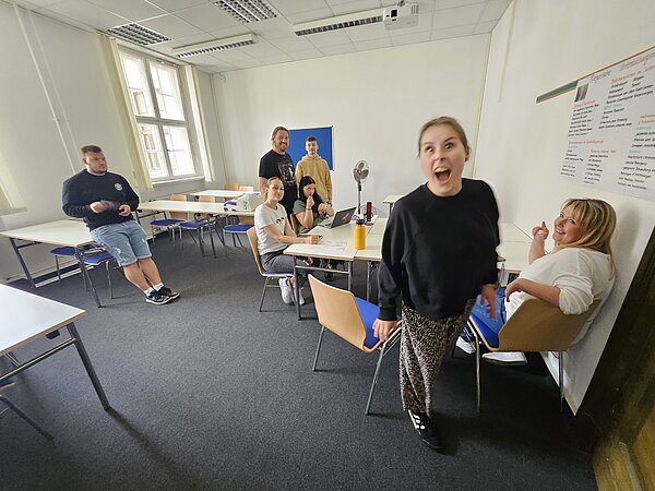 Podcast-Projekt: KAFFEE TRINKEN & SPIELEN Vorne steht eine Schülerin mit schwarzen Klamotten, die in die Kamera schaut, dahinter sitzen weitere Schülerinnen.