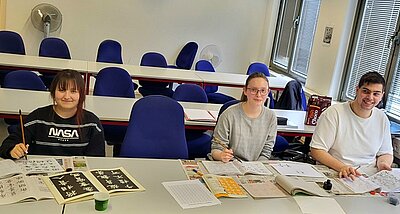 Zwei Schülerinnen und ein Schüler der Euro Akademie Hannover sitzen im Unterrichtsraum, haben Übungshefe zu Kalligrafie, die nötigen Schreibutensilien vor sich und lächeln für ein gemeinsames Foto in die Kamera.