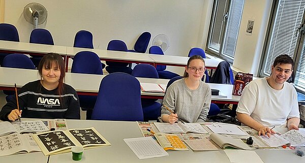 Chinesische Kalligrafie –Vier Schätze des Gelehrtenzimmers Zwei Schülerinnen und ein Schüler der Euro Akademie Hannover sitzen im Unterrichtsraum, haben Übungshefe zu Kalligrafie, die nötigen Schreibutensilien vor sich und lächeln für ein gemeinsames Foto in die Kamera.
