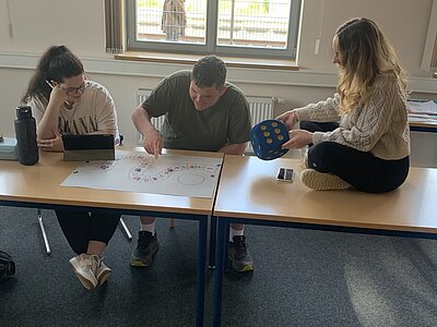 Drei Personen sitzen an einem Tisch und spielen ein Brettspiel. Eine Person hält einen großen blauen Würfel, während die anderen beiden sich auf das Spielbrett konzentrieren. Auf dem Tisch liegen Spielsteine und Karten.
