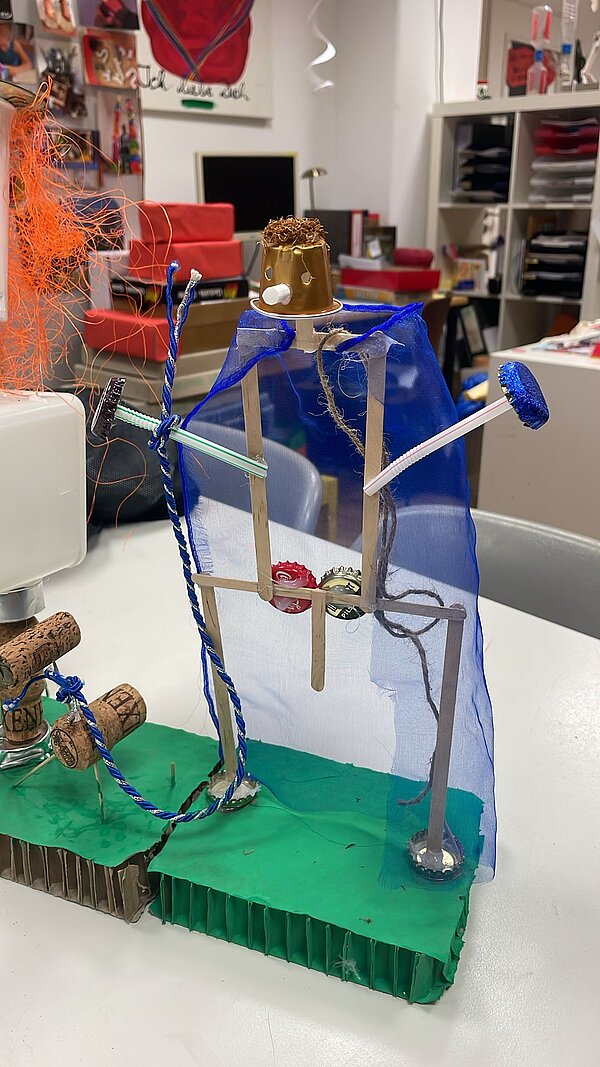 Eine aus Eisstielen zusammengeklebte Figur mit Strohhalmen als Arme und einem blauen Umhang steht auf grüner Pappe.