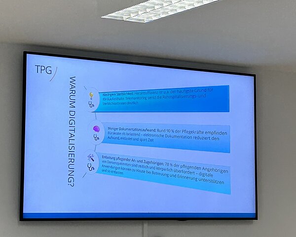 Eine Präsentationsfolie auf einem Wandbildschirm zeigt eine Infografik mit dem Titel 'WARUM DIGITALISIERUNG?'. Links steht das Logo 'TPG'. Die Grafik enthält drei farbige Balken mit Symbolen und kurzen Texten. Der erste Balken ist blau und enthält ein Glühbirnensymbol. Der zweite Balken ist violett und zeigt ein Dokumentensymbol. Der dritte Balken ist türkis und enthält ein Symbol mit zwei Personen. Die Balken sind jeweils mit kurzen, gut lesbaren Texten versehen, die Argumente für die Digitalisierung im Pflegebereich nennen. Die Decke mit einer weißen Rasterleuchte ist im oberen Bildbereich sichtbar.
