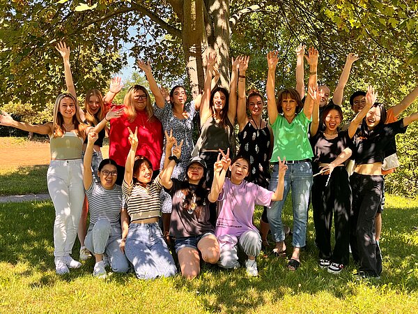 Herzlichen Glückwunsch! Eine Gruppe von 14 fröhlichen Menschen, hauptsächlich Frauen, posiert unter einem Baum auf einer grünen Wiese. Sie heben die Arme in die Luft und lachen. Einige zeigen das Peace-Zeichen, andere strecken ihre Hände in die Höhe. Die Gruppe ist bunt gekleidet, mit verschiedenen Outfits von leger bis sommerlich schick. Die Sonne scheint, und der Hintergrund zeigt Natur mit Bäumen und Wiese.