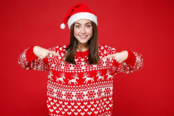 Lächelnde junge und gut gelaunte Frau mit rot-weißem Weihnachtspullover und Weihnachtsmütze, die mit den Fingern halb nach unten zeigt, der Hintergrund ist rot.