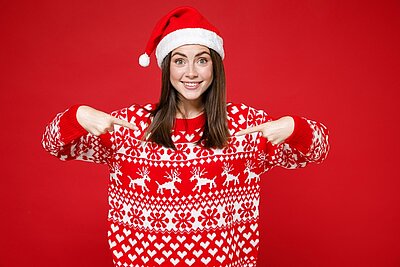 Lächelnde junge und gut gelaunte Frau mit rot-weißem Weihnachtspullover und Weihnachtsmütze, die mit den Fingern halb nach unten zeigt, der Hintergrund ist rot.