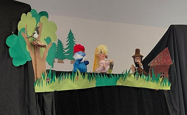 Bühnenbild mit Handpuppen vor einem Hintergrund aus grünem Papier, das Gras, Bäume und einen Zaun darstellt. Links sitzt ein Vogel in einem Nest auf einem Baum. Daneben steht eine blaue Figur mit roter Mütze, gefolgt von einer blonden Puppe mit rosa Kleid. Rechts steht eine männliche Puppe mit braunem Hut und Bart vor einem kleinen Haus mit rotem Dach. Schwarzer Stoff bildet den Bühnenvorhang.