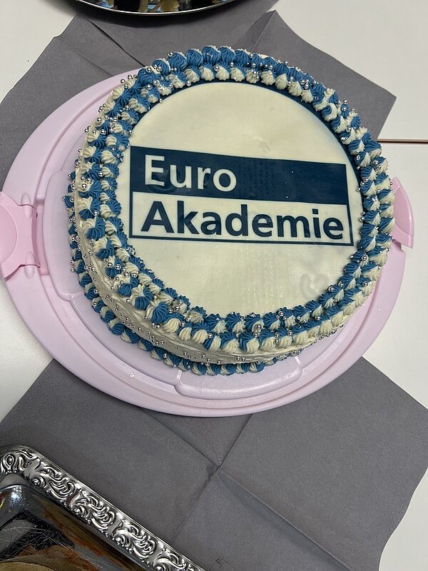 Runde Torte mit weißer Glasur, verziert mit zwei Reihen aus blauen und weißen Cremetupfen am Rand. In der Mitte befindet sich eine glatte Fläche mit dem rechteckigen Logo der Euro Akademie, das aus einem blauen Balken und dem Schriftzug 'Euro Akademie' besteht. Die Torte steht auf einer rosa Kunststoffplatte, darunter liegt eine graue Serviette.