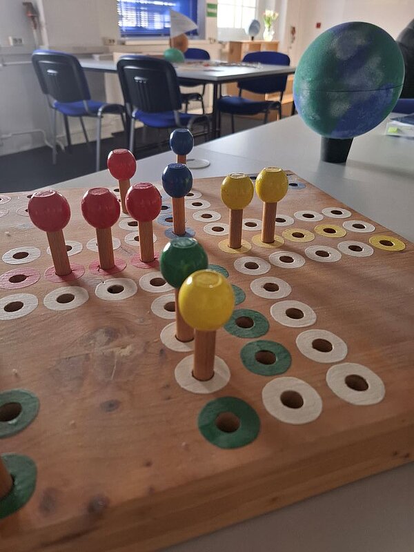 Ein Brettspiel mit farbigen Knöpfen und Ringen auf einer Holzplatte. Die Knöpfe und Ringe sind in verschiedenen Farben wie Rot, Gelb, Grün und Blau angeordnet. Im Hintergrund sind Stühle und ein Tisch zu erkennen.