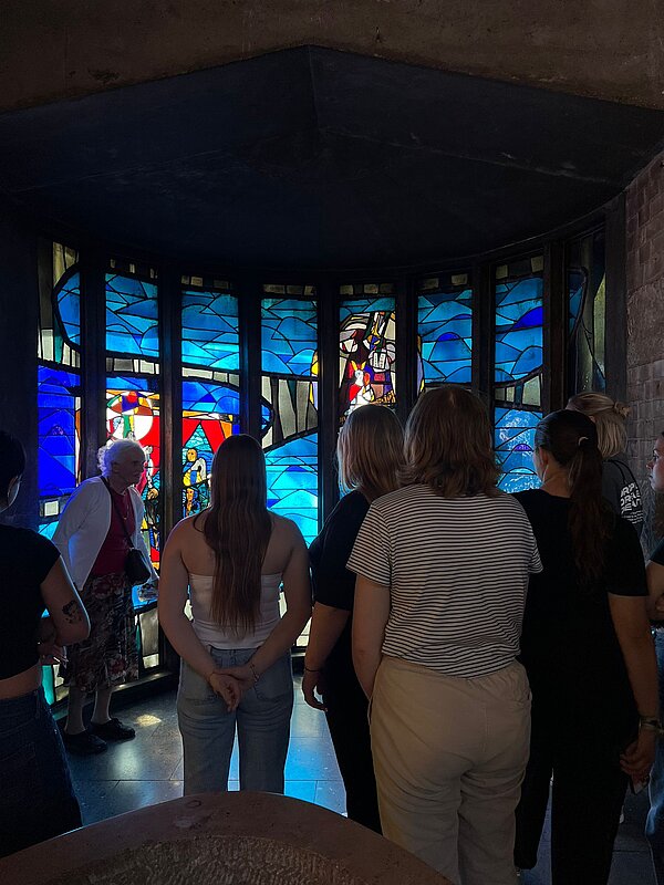 Besuchergruppe betrachtet farbenfrohes Kirchenfenster mit blauen und roten Glasmalereien im Inneren einer Kirche, vorne ist ein steinerner Taufstein sichtbar.