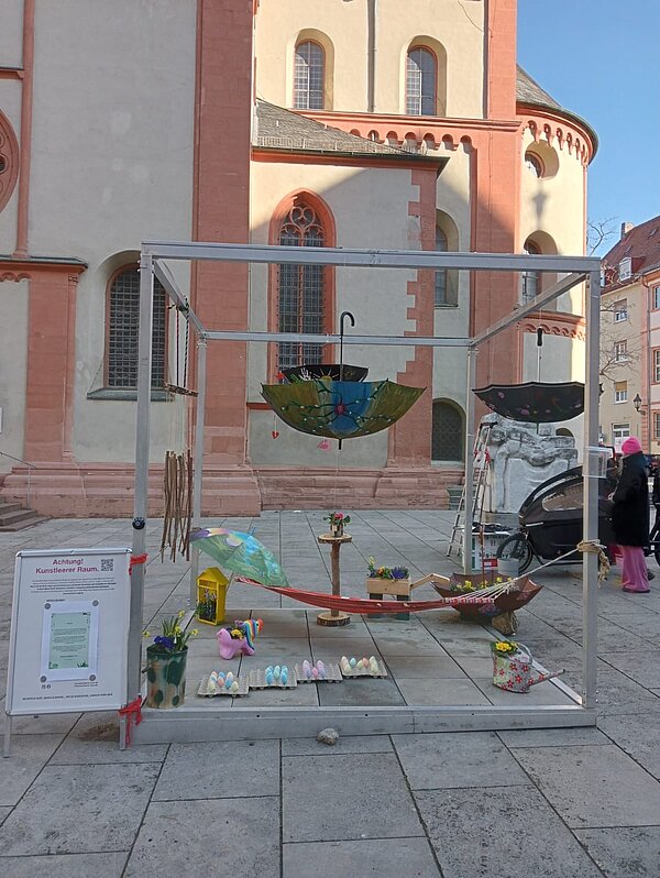 Vor dem Würzburger Dom sind innerhalb eines Metallrahmens verschiedene Kunstobjekte ausgestellt, darunter mehrere Regenschirme und eine Hängematte.