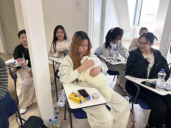 Mehrere Personen sitzen in einem Klassenraum auf Stühlen mit kleinen, angebauten Tischen. Eine Person in der Mitte hält ein weißes, flauschiges Stofftier im Arm. Auf den Tischen liegen verschiedene Gegenstände wie Wasserflaschen, Snacks, Schreibutensilien und Mobiltelefone. Im Hintergrund ist ein geöffnetes Fenster zu sehen, durch das Tageslicht einfällt.