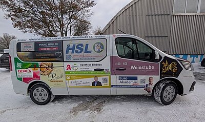 Ein weißer Transporter steht auf schneebedecktem Boden vor einer Industriehalle. Die Fahrzeugseite ist mit zahlreichen, farbigen Werbeflächen beklebt. Im unteren Bereich befindet sich ein rechteckiges, blau-weißes Werbebanner der Euro Akademie. Das Logo und der Schriftzug 'Euro Akademie' sind deutlich zu erkennen. Daneben ist ein Porträtfoto einer Person abgebildet. Weitere Werbeflächen auf dem Fahrzeug zeigen Logos, Slogans und Kontaktdaten verschiedener regionaler Unternehmen. Im Hintergrund sind kahle Bäume und ein Metallgebäude zu sehen.