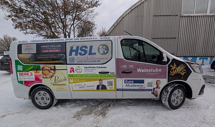 Ein weißer Transporter steht auf schneebedecktem Boden vor einer Industriehalle. Die Fahrzeugseite ist mit zahlreichen, farbigen Werbeflächen beklebt. Im unteren Bereich befindet sich ein rechteckiges, blau-weißes Werbebanner der Euro Akademie. Das Logo und der Schriftzug 'Euro Akademie' sind deutlich zu erkennen. Daneben ist ein Porträtfoto einer Person abgebildet. Weitere Werbeflächen auf dem Fahrzeug zeigen Logos, Slogans und Kontaktdaten verschiedener regionaler Unternehmen. Im Hintergrund sind kahle Bäume und ein Metallgebäude zu sehen.