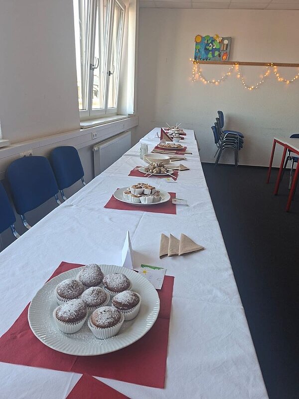 Ein langer Tisch ist mit verschiedenen Platten mit Gebäck und Snacks gedeckt. Auf dem Tisch sind auch kleine Zettel und Servietten zu sehen. Seitlich befinden sich Stühle und ein Fenster. Der Raum ist mit einer Lichterkette dekoriert.