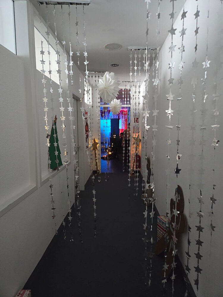 Schmaler, weißer Flur, der mit herunterhängenden silbernen Stern-Girlanden und großen weißen Papiersternen dekoriert ist; am Ende leuchtet ein bunt geschmückter Raum im Hintergrund.