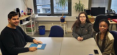 Bitte lächeln Zwei weibliche und ein männlicher Schüler*innen sitzen gemeinsam an einem großen weißen Tisch und lächeln für ein Foto in die Kamera.