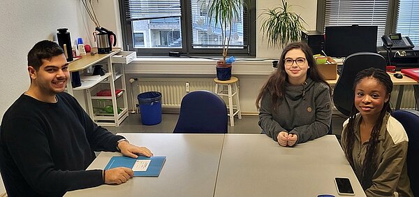 Erasmus-Speed-Dating Zwei weibliche und ein männlicher Schüler*innen sitzen gemeinsam an einem großen weißen Tisch und lächeln für ein Foto in die Kamera.