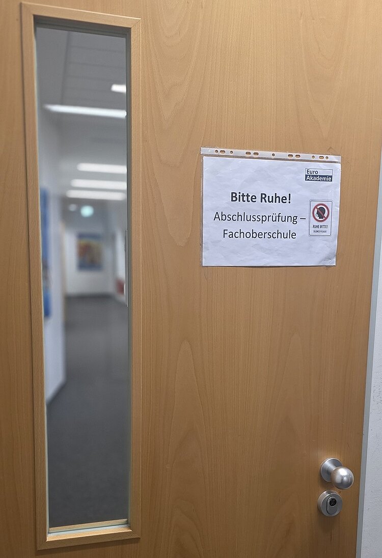 Holztür mit schmalem, senkrechtem Sichtfenster. An der Tür ist ein weißes Blatt Papier befestigt, darauf steht in klar lesbarer Schrift: 'Bitte Ruhe! Abschlussprüfung – Fachoberschule'. Daneben befindet sich ein rundes Symbol mit durchgestrichenem Handy. Durch das Fenster ist ein leerer, heller Flur mit weißen Wänden und mehreren Türen zu sehen.