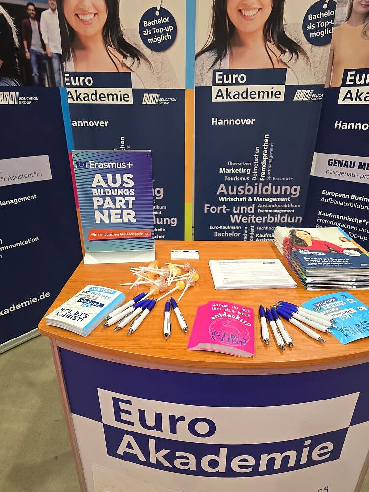 Ein Ausschnitt eines Infotischs der Euro Akademie Hannover. Auf dem Holztisch liegen mehrere Stifte, ein Stapel Broschüren mit dem Titel „AUS BILDUNG PARTNER“ und ein pinkfarbenes Notizbuch mit der Aufschrift „WEIL DU’S KANNST!“. Im Hintergrund sind weitere Roll-up-Banner der Euro Akademie mit Informationen zu Ausbildungsmöglichkeiten und Studiengängen sichtbar. Ein Banner zeigt eine lächelnde Person und wirbt für den „Bachelor als Top-up möglich“.