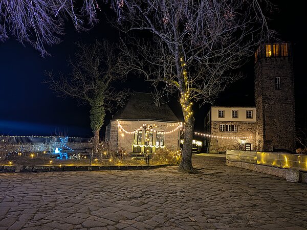 Nächtlicher Burghof mit Kopfsteinpflaster, Bäumen und warmen Lichterketten, kleine historische Gebäude und Turm im Hintergrund.