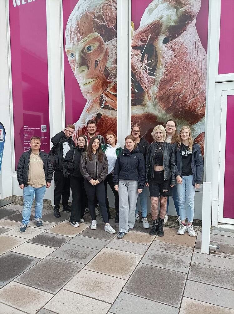 Eine Gruppe junger Menschen steht lächelnd vor dem Eingang zur Ausstellung "Körperwelten". Hinter ihnen ist ein großes, pinkfarbenes Plakat mit einer plastinierten menschlichen Figur zu sehen. Der Boden ist gepflastert und wirkt leicht feucht.