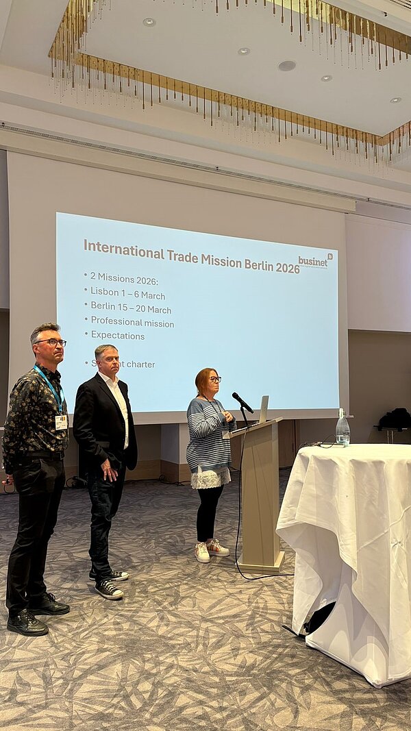 Drei Personen stehen in einem Konferenzraum vor einer Leinwand. Eine Frau spricht an einem Rednerpult mit Mikrofon, während zwei Männer daneben stehen. Die Leinwand zeigt eine Präsentationsfolie mit dem Titel 'International Trade Mission Berlin 2025' und mehreren Aufzählungspunkten auf Englisch. Im Vordergrund steht ein runder Tisch mit weißer Tischdecke. Der Raum hat hohe Decken und moderne Beleuchtung.