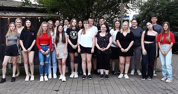 Herzlich willkommen Eine Gruppe von Jugendlichen posiert für ein Gruppenfoto. Die Mehrzahl sind Mädchen, es sind nur drei Jungen auf dem Bild. Die Personen tragen sommerliche Kleidung und stehen in einem Hof vor einem Baum.