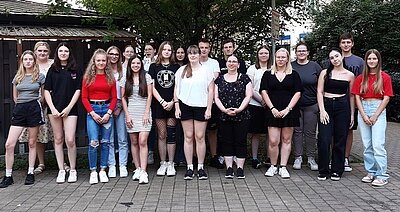 Eine Gruppe von Jugendlichen posiert für ein Gruppenfoto. Die Mehrzahl sind Mädchen, es sind nur drei Jungen auf dem Bild. Die Personen tragen sommerliche Kleidung und stehen in einem Hof vor einem Baum.