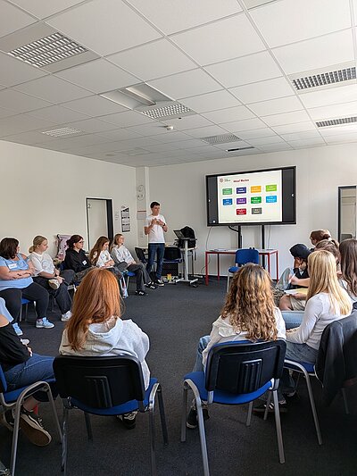 Die Gruppe der Auszubildenden sitzt in einem Seminarraum und hört einem Vortragenden zu. Auf einem großen Bildschirm ist eine farbige Präsentationsfolie zum Thema Abfall zu sehen.
