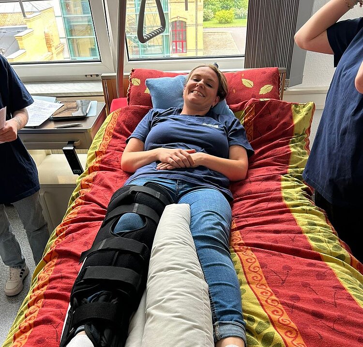 Eine lächelnde Person liegt in einem Krankenhausbett mit einer Beinschiene am rechten Bein. Sie trägt ein blaues T-Shirt und Jeans. Neben dem Bett stehen zwei Personen in blauen T-Shirts, eine davon hält ein Dokument. Der Raum ist hell und hat große Fenster.