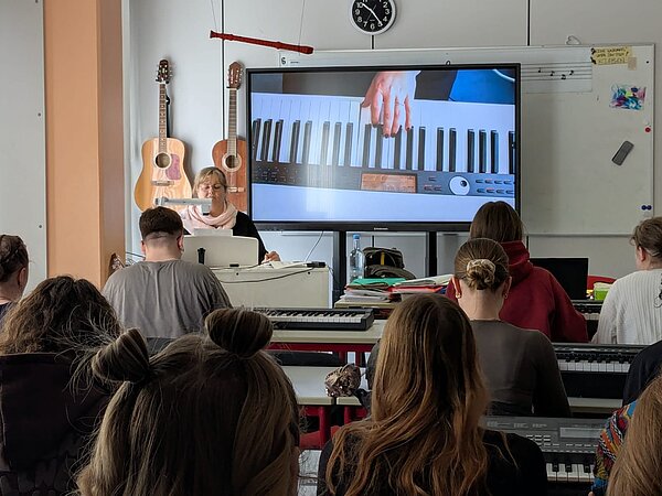 In einem Klassenzimmer sitzen drei Reihen Schüler vor einer Lehrerin und einer elektronischen Tafel, auf der eine Hand auf einer Klaviertastatur zu sehen ist. An der Wand daneben hängen zwei Gitarren.