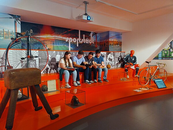 Mehrere Jugendliche sitzen in einer Ausstellung zum Thema Sportgeschichte auf einem roten Podest. Um sie herum sind historische Sportgeräte wie ein Hochrad, ein alter Turnbock, Sportkleidung und ein Fahrrad ausgestellt.