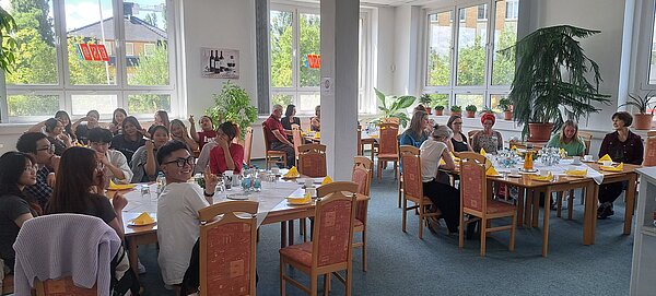 Speed-Dating für Pflegefachkräfte: Eine gelungene Vorbereitung für die Ausbildung Zu sehen ist ein großer Raum Raum mit einer weißen Säule in der Mitte. In dem Raum stehen überall in Vierecke zusammengestellte Tische mit Stühlen, auf denen jüngere mit mittelalte Personen sitzen.