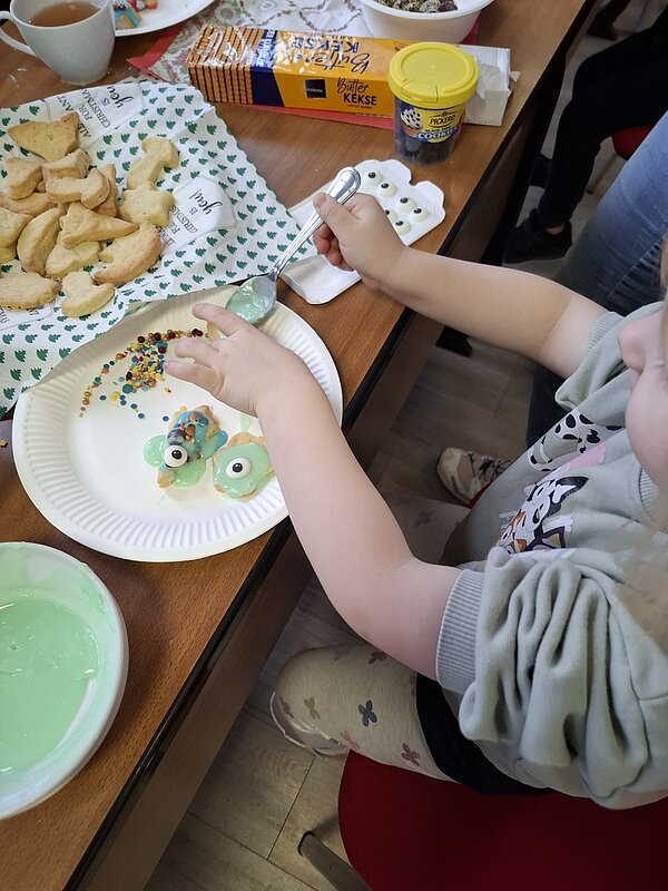 Ein Kind sitzt am Tisch und verziert Kekse mit grünem Zuckerguss und bunten Streuseln, umgeben von weiteren Backzutaten
