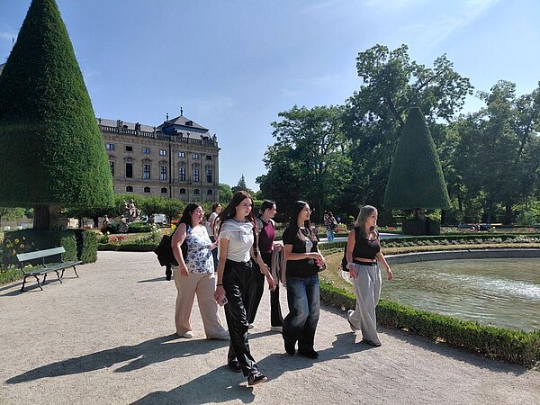 Eine Gruppe von Personen spaziert auf einem Weg im Hofgarten der Würzburger Residenz. Im Hintergrund sind das Gebäude der Residenz, ein Gewässer und die grünen Hecken des Gartens zu sehen.