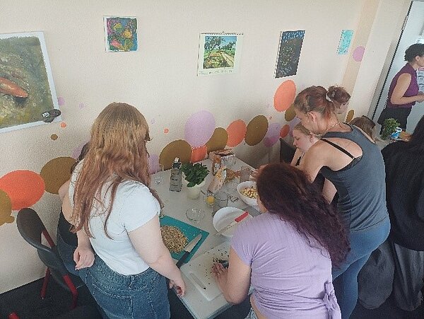 Ein gesunder Abschluss Schülerinnen und Schüler stehen um einen Tisch herum, auf dem Brettchen und Lebensmittel stehen und bereiten diese zu.