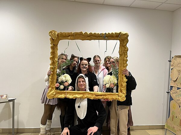 Sieben Studierende schauen lächelnd in ihren Faschingskostümen durch einen übergroßen selbst gestalteten Bilderrahmen und posieren für ein Foto.