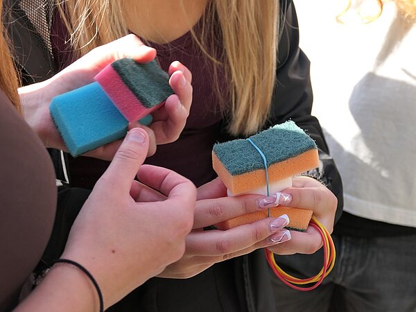 Mehrere Hände halten bunte Haushaltsschwämme. Ein Schwamm ist mit einem Gummiband umwickelt, daneben liegen weitere Gummibänder. Die Fingernägel einer Person sind lang und lackiert. Die Szene spielt im Freien bei Tageslicht.