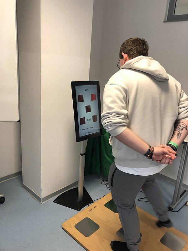 Ereignisreicher Tag für die angehenden Erzieher*innen Ein Schüler mit weißem Kapuzenpulli steht auf einer Art Simulator. Er trägt nur Strümpfe und springt auf einem Brett, das am Boden liegt und in verschiedene Bereiche eingeteilt ist, von einem Bereich zum anderen, und schaut dabei auf einen Bildschirm.