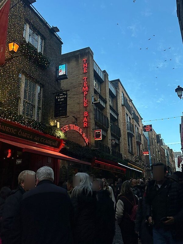 Belebte Straße in Dublin mit dem berühmten Pub 'The Temple Bar'. Das Gebäude ist mit roten Neonleuchten beschriftet, und viele Menschen sind auf der Straße unterwegs. Im Hintergrund sieht man weitere historische Gebäude und eine blassblaue Abendhimmel.